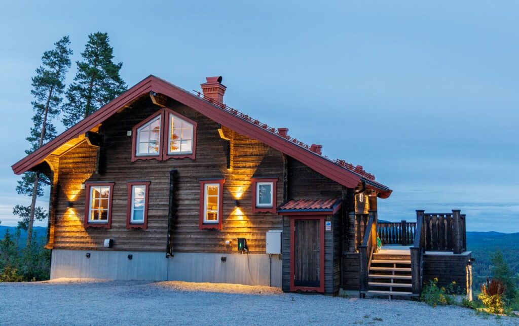 A cozy wooden cabin lit up at dusk, nestled in a serene rural landscape with mountain views.